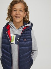 Námornícky modrý prešívaný vest s chevronovým prešívaním, červenými akcentmi a logom. Nosený cez šedú mikinu s potlačou.