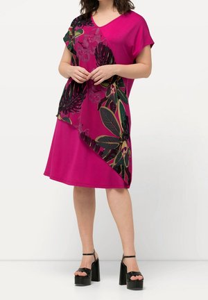 Fuksjowa sukienka z krótkimi rękawami, dekoltem w kształcie litery V i kwiatowym wzorem w kolorze purpurowym, zielonym i czarnym. Asymetryczny dół o gładkiej fakturze. Czarne sandały na platformie.