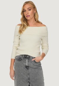 Off-white off-the-shoulder ribbed sweater with long sleeves, paired with a gray denim skirt featuring a front slit and embellishments.