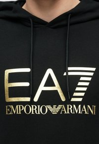Fekete kapucnis pulóver arany logóval, rajta az "EA7 EMPORIO ARMANI" felirat. Cipzáras kapucnival és puha anyaggal készült. Alkalmi viseletre tervezve.