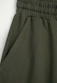 Ciemnozielone dresowe spodnie z elastycznym pasem i widoczną boczną kieszenią na białym tle.