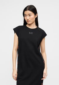 Γυναίκα με μακριά σκουρόχρωμα μαλλιά φοράει χαλαρό μαύρο φόρεμα με κοντά μανίκια και λογότυπο EA7 στο στήθος και στις πλευρές, στέκεται μπροστά σε απλό φόντο.