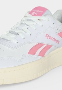 Λευκό αθλητικό παπούτσι Reebok με ανοιχτό ροζ λογότυπο και ρίγες, λευκά κορδόνια και μπεζ σόλα, εμφανίζεται σε κοντινό πλάνο από το πλάι και από πάνω.