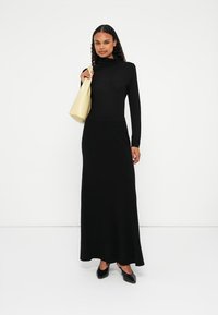 Abito nero a maniche lunghe con scollo a dolce vita, parte superiore aderente e gonna svasata, tessuto morbido. Abbinate a una borsa gialla e tacchi neri.