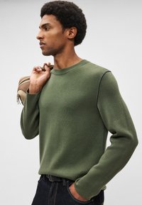 Camisola verde oliva com mangas compridas, gola canelada e costura escura contrastante ao longo do ombro. Textura lisa, ajuste casual.