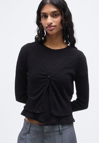 Μαύρη ριμπ ζακέτα με στρογγυλή λαιμόκοψη, κλείσιμο με κουμπιά στο μπροστινό μέρος και κυματιστές άκρες. Μακριά μανίκια και ελαφρώς φαρδύ τελείωμα.