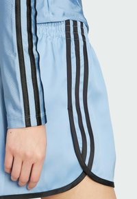 Ljusblå idrottsshorts med svart elastisk midja. Tre svarta ränder går vertikalt längs sidorna. Slät, glänsande tygtextur.