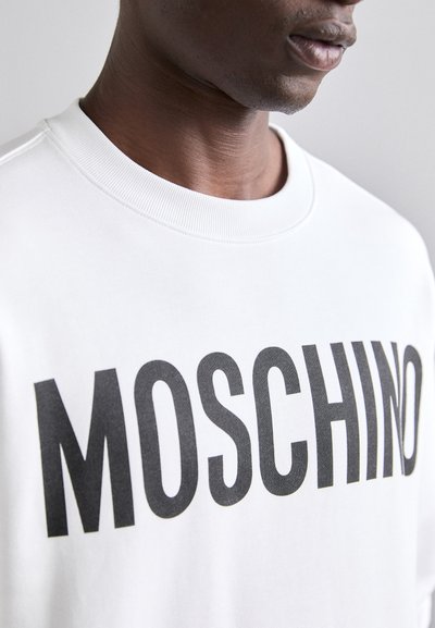 Sudadera blanca con "MOSCHINO" en letras grandes de color negro. La tela parece suave, con un cuello redondo y detalle de cuello acanalado.