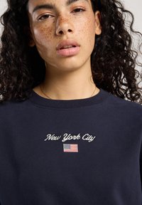 Joven con cabello rizado y pecas vistiendo una sudadera azul marino bordada con "New York City" y una pequeña bandera de Estados Unidos.