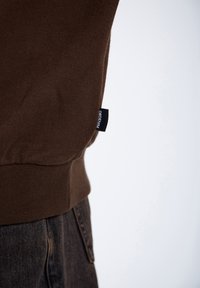 Felpa marrone in cotone con orlo a costine, etichetta laterale con la scritta "DRDENIM," abbinata a jeans in denim scuro.