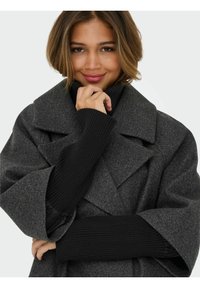 Szary płaszcz z mieszanki wełny z szerokim kołnierzem i dużymi klapami, noszony na czarnym prążkowanym golfie. Charakteryzuje się luźnymi rękawami i strukturalnym wykończeniem.