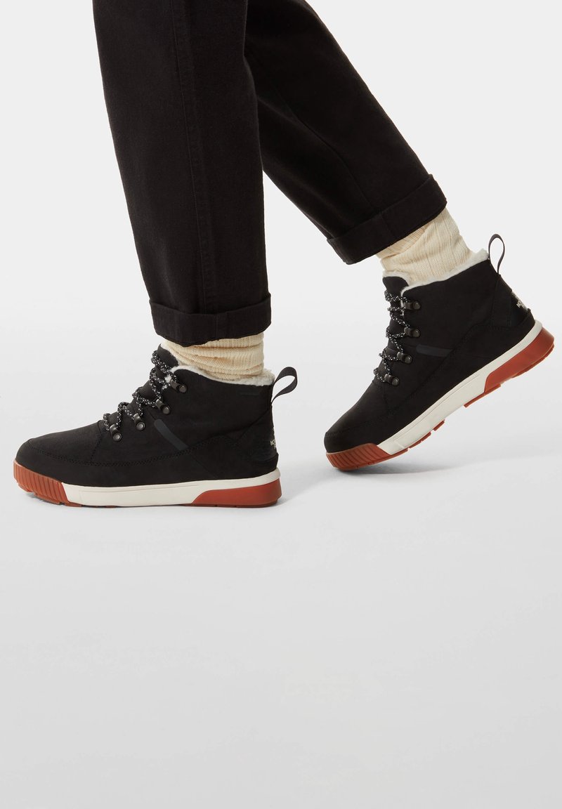 Personne portant des baskets montantes noires avec des semelles blanches et orange, des chaussettes crème et un pantalon noir retroussé, posant le pied sur une surface blanche.