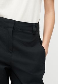 Άτομο που φοράει μαύρο παντελόνι σε ραφή, με το χέρι στην τσέπη και λευκό πουκάμισο μέσα στη ζώνη, εμφανιζόμενο από τη μέση έως το μέσο του μηρού.