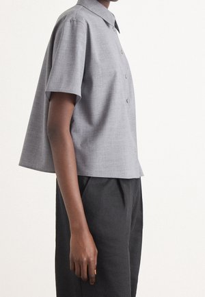 Osoba ubrana w szarą koszulę na guziki z krótkim rękawem i czarne spodnie, widziana z boku na jednolitym białym tle.