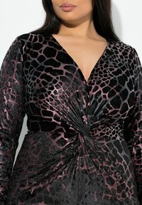 Musta ja viininpunainen mekko, jossa on V-kaula-aukko, eläinkuosilla varustettu muotoilu, kierretty yksityiskohta vyötäröllä ja pitkät hihat, joissa on teksturoitu pinta.