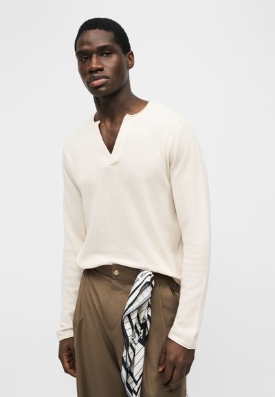 Άνδρας που φοράει κρεμ μακρυμάνικη μπλούζα με V λαιμόκοψη και καφέ παντελόνι με ένα μαύρο-λευκό φουλάρι με σχέδιο που είναι κρυμμένο στη μέση.