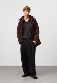 Giacca a vento bordeaux con cappuccio sopra un maglione scuro a scollo a V, abbinata a pantaloni neri plissettati e scarpe nere senza lacci.