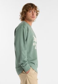 Felpa verde realizzata in materiale morbido, con maniche lunghe, polsini a costine e una stampa del logo bianco sulla parte frontale. Abbinata a pantaloni beige.