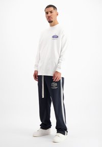 Wit overhemd met lange mouwen en grafische accenten, gecombineerd met marineblauwe sweatpants met witte strepen en logo, afgewerkt met witte sneakers.