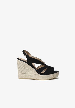 Sandale en suede noire à talon compensé avec un design de brides entrecroisées. Caractérisée par un talon enveloppé de corde naturelle texturée et un semelle intérieure lisse.