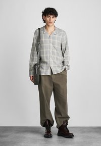 Grijs geruit overhemd met knopen, losse beige broek, bruine veterlaarzen. Zwarte schoudertas met verstelbare band. Eenvoudig, functioneel ontwerp.