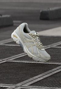 Light gray athletic shoe with a mesh upper, textured overlays, and a cushioned sole. Features beige accents and a slightly reinforced toe.