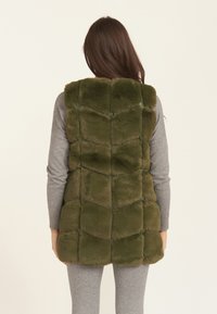 Green faux fur vest with a quilted pattern, featuring a high collar and a soft texture. Paired with a gray long-sleeve top underneath.