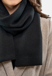 Sort strikkscarf med en glat overflade og ribbede ender, draperet over en blød, beige frakke. Har en medium tykkelse og et hyggeligt design.