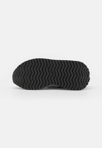 Suela de zapato de goma negra con patrón de banda de rodadura en zigzag y el texto "new balance" repetido sobre un fondo blanco liso.