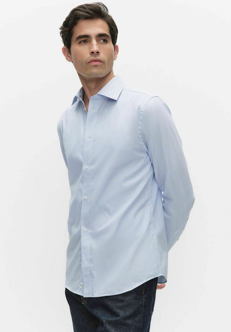 Młody mężczyzna ubrany w jasnoniebieską koszulę w kratę oraz ciemne dżinsy, stojący z rękami za plecami, patrzący w lewo.