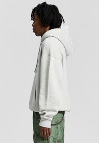 Hoodie oversized grigio chiaro con cappuccio regolabile, polsini a coste e tasca a marsupio, indossato con pantaloni verdi caratterizzati da un dettaglio a catena.
