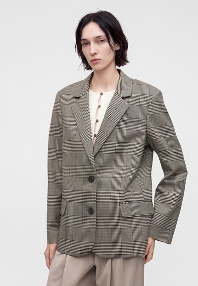 Femme aux cheveux courts et foncés porte un blazer marron à carreaux par-dessus un haut blanc boutonné et un pantalon beige ample sur un fond uni.
