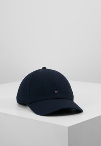 Căciulă albastră închis cu o vizieră curbată, realizată din bumbac. Dispune de un mic logo roșu, alb și albastru pe față. Design clasic de șapcă de baseball.