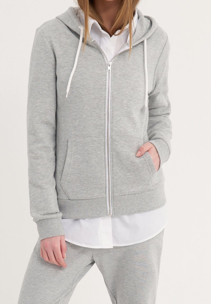 Personne portant un sweat à capuche gris clair zippé avec des cordons blancs, associé à un pantalon de survêtement assorti et un t-shirt blanc en dessous.