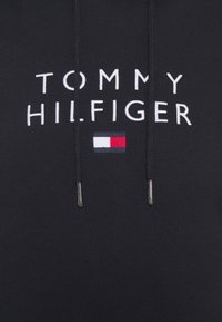 Sudadera negra con texto bordado en blanco "TOMMY HILFIGER" y un pequeño parche de logo en rojo, blanco y azul con extremos de cordón plateados.