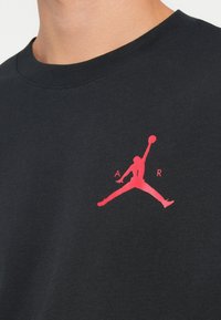 Fekete pamut póló rövid ujjú fazonnal, amelyen egy rózsaszín Air Jordan logó található a mellrészen. Egyszerű, klasszikus kerek nyakkivágás.