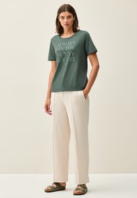 T-shirt vert olive avec un graphisme textuel, associé à un pantalon large crème et des sandales vertes. Tissu doux et coupe décontractée.