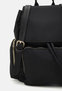 Sac à dos en nylon noir avec détails en cuir, comprenant une poche avant zippée, des bretelles réglables et des éléments en métal doré.