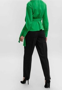 Blusa verde in satin con vita a peplum e maniche lunghe, abbinata a pantaloni neri e scarpe nere con tacco alto. Tessuto liscio, design elegante.