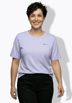 Persona sorridente con capelli corti e ricci che indossa una maglietta sportiva viola chiaro e pantaloni neri su sfondo bianco.