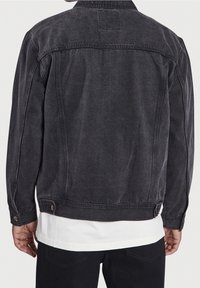 Hombre con una chaqueta de mezclilla negra desgastada sobre una camisa blanca y pantalones negros, mostrado de espaldas frente a un fondo liso.