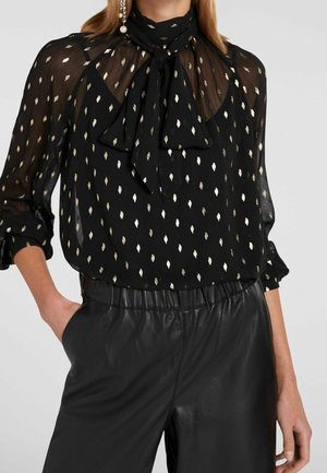 Schwarze transparente Bluse mit goldenem Diamantmuster, Schleifenkragen, hochgekrempelten langen Ärmeln, kombiniert mit schwarzen, hoch taillierten Lederhosen.