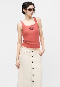 Tânără purtând ochelari de soare, un top roșu fără mâneci cu un logo și o fustă bej cu talie înaltă și nasturi, stând în fața unui fundal simplu și deschis la culoare.