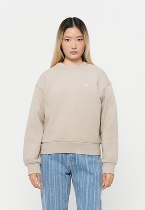 Beige genser med lange ermer, ribbede mansjetter og kant. Har et lite logo på brystet. Kombinert med lyseblå stripete jeans.