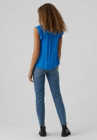 Donna con capelli ricci che indossa un blusa senza maniche di un brillante blu, jeans blu slim-fit e scarpe nere, in piedi con le spalle rivolte verso di noi su uno sfondo neutro.