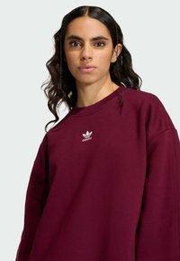 Burgundská mikina s kulatým výstřihem, spadlými rameny a bílým logem Adidas na hrudi. Materiál z bavlny, volný střih.