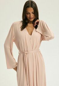 Jasnoróżowy szlafrok z długimi rękawami, dekoltem w szpic, paskiem w talii oraz miękkim, lekko opadającym materiałem z delikatnymi fałdami.
