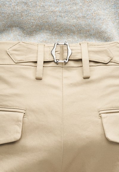 Μπεζ παντελόνι με ζώνη που έχει ασημένια μεταλλική αγκράφα, συνδυασμένο με πλεκτό πουλόβερ σε πολλαπλές αποχρώσεις του μπεζ.