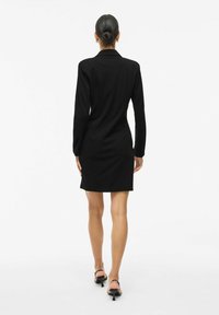 Schwarzes Langarmkleid mit Kragen, tailliertem Schnitt und Länge bis zur Mitte des Oberschenkels. Glatter Stoff mit minimalen Details, kombiniert mit schwarzen Absatzsandalen.