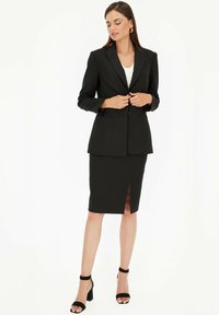 Czarna, dopasowana marynarka na białej bluzce, zestawiona z ołówkową spódnicą do kolan z rozcięciem z przodu. Modelka nosi czarne sandały na słupku.